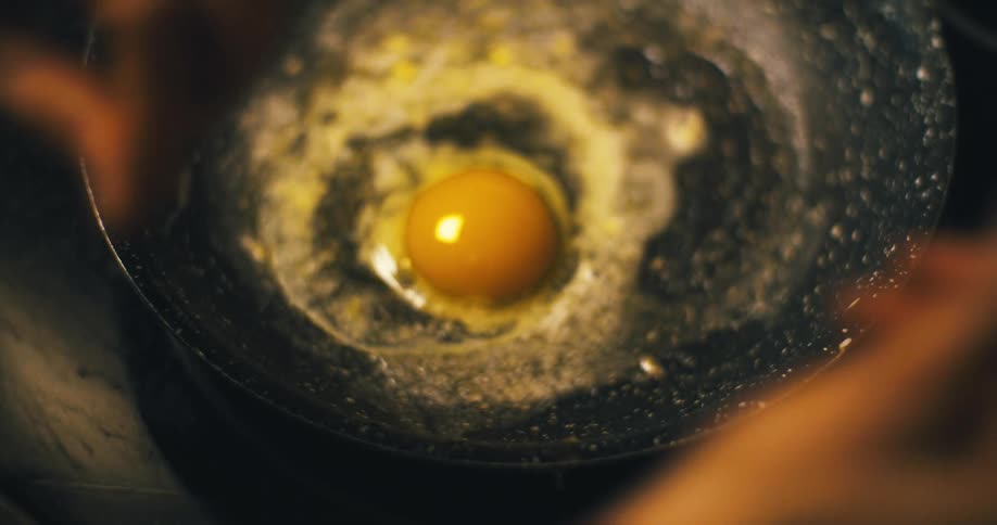 A close view of a person cracking an egg into a skillet shows the yolk and egg white cooking and sizzling