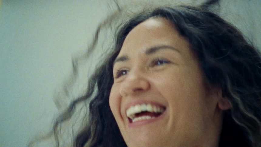 The video starts with a shot of a bed with a white sheet on it followed by a sudden reveal of a person with long dark curly hair laughing the video ends with a closeup on the persons face as they smile
