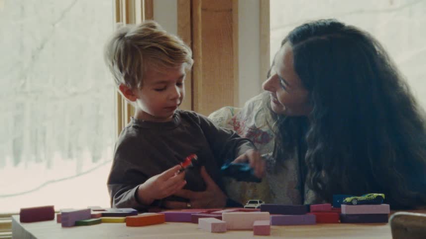 A woman with dark hair sits with a child with light colored hair as they both play with toy cars and building blocks on a wooden table