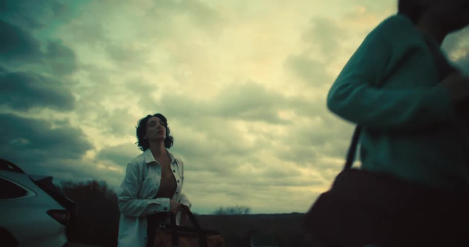 A woman and a girl walk near an open car trunk holding large brown bags with a cloudy sky behind them
