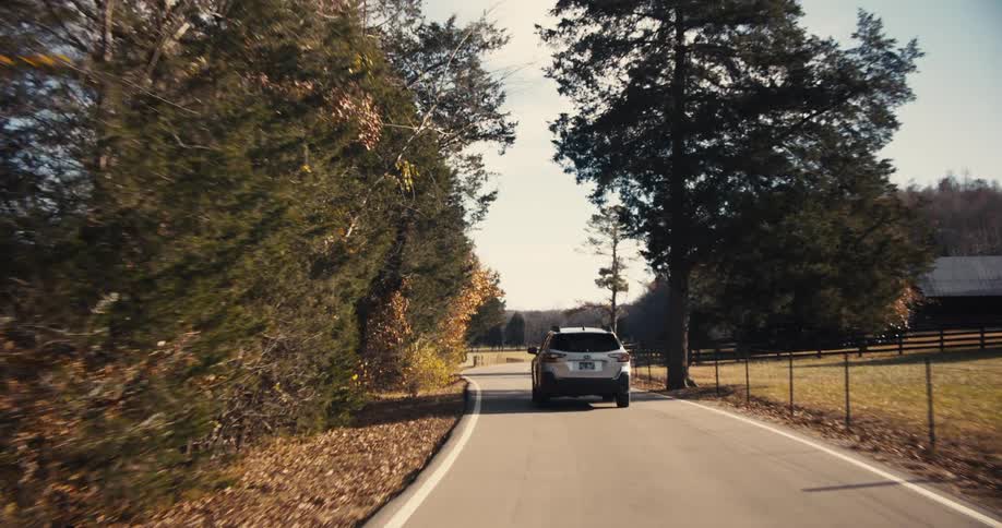 A white suv is driving away from the camera on a twolane country road with a wooded area on one side and a fence and field on the other