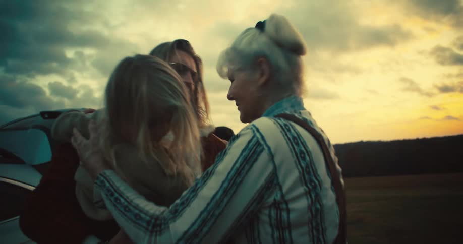 The video shows a group of people appearing to be a family standing around a car at sunset a man with long blonde hair is holding a child and an older woman walks up to greet them with a hug
