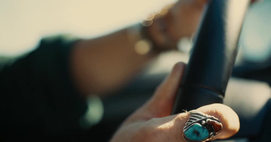A hand grips a steering wheel adorned with a large turquoise and silver statement ring