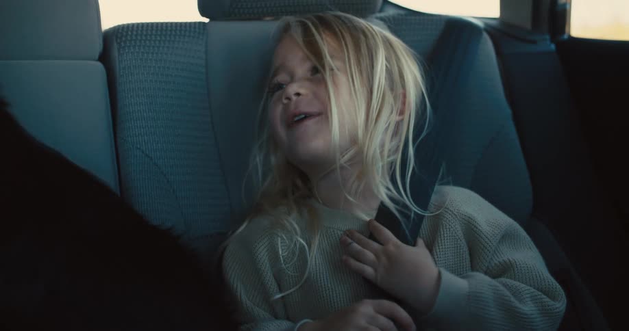 A child with blonde hair wearing a beige sweater is sitting in the back seat of a car fidgeting with a seatbelt while looking away from the camera
