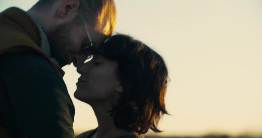 A closeup profile shot of a man and a woman about to kiss with the man in a tan vest and glasses looking down at the woman as the woman looks up at him with a loving expression