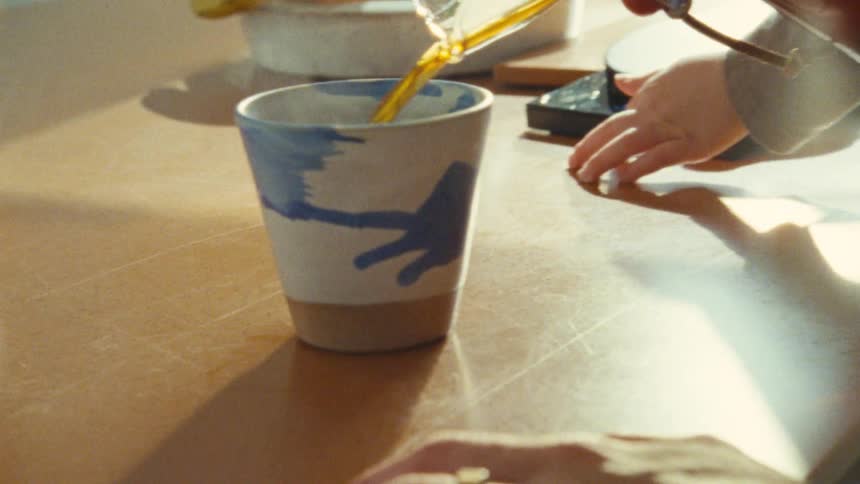 The video shows a closeup of a person pouring a liquid from a small clear container into a blue and white patterned ceramic mug on a table the background is a brightly lit kitchen setting