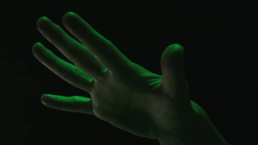 A close view of a human hand in a dark space illuminated by a green light as the fingers close and unclose into a fist