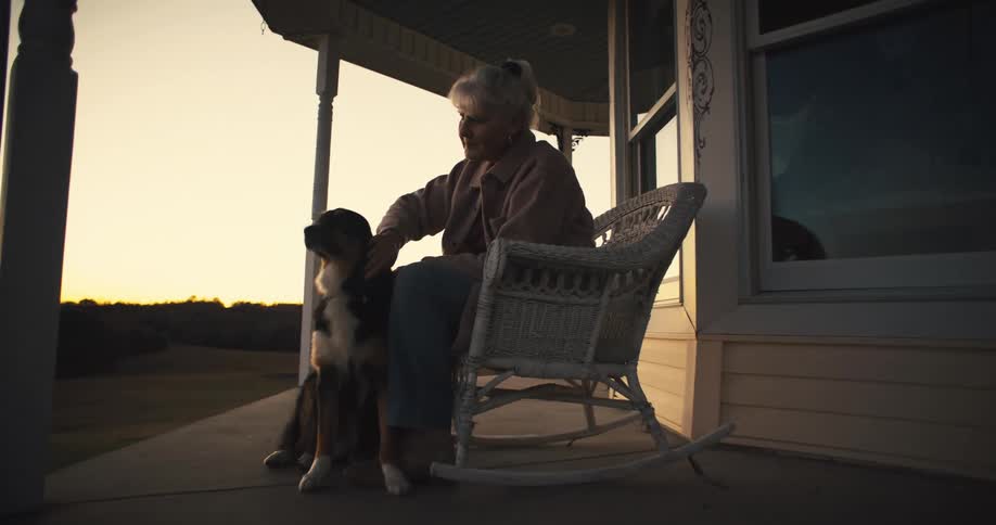 Elderly woman pets her dog on a porch at sunset