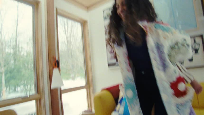 A woman with long dark hair is seen holding a child with short light colored hair on her back as they both smile and she dances on a brightly lit living room with framed art and a yellow couch