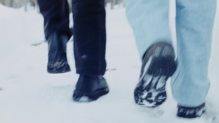 A shot from behind and below of two people walking together on a snowy ground with a focus on their feet and shoes