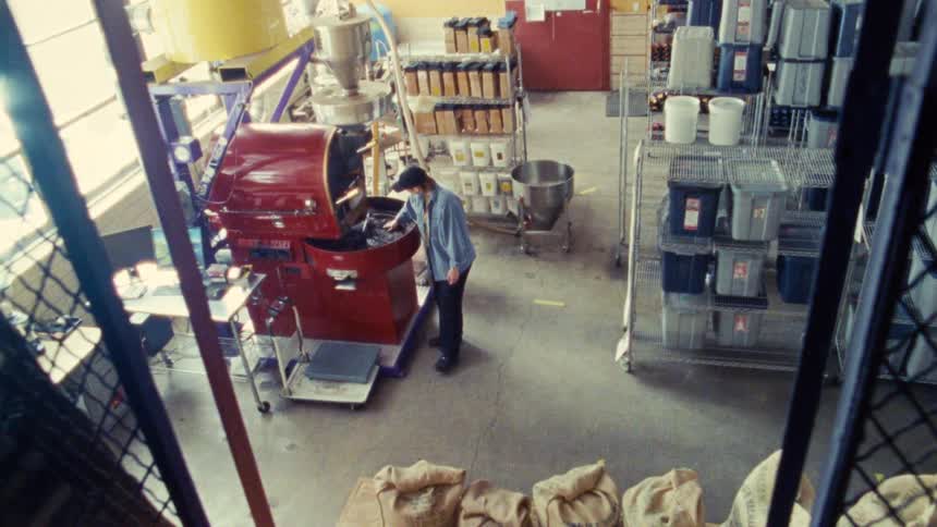Based on the video a wide shot from a high angle shows a person in a blue shirt walking past a red coffee roasting machine in what appears to be a warehouse or factory the area is filled with bags of beans and metal shelving units