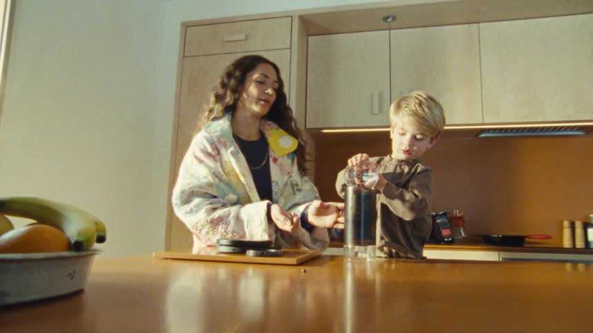 The video shows a woman with long dark hair and a young child with blonde hair standing at a kitchen counter they are looking at a coffee grinder on the counter