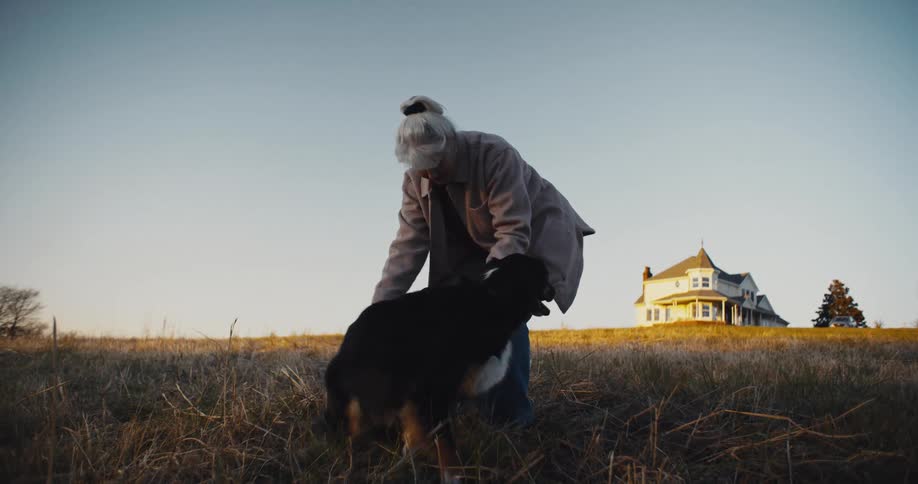 Elderly woman pets a dog in an open field with a large house in the background