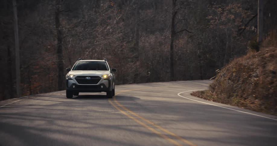 A silver car driving along a rural road with trees and hills visible on either side under a cloudy sky