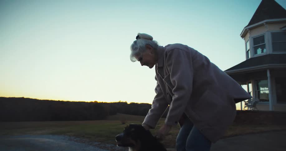 An elderly woman holding a dog in front of a house while a car drives away