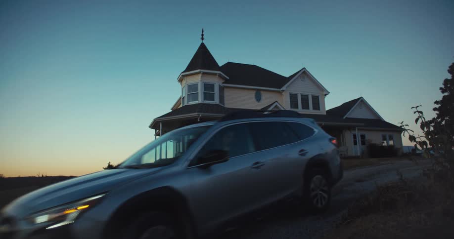 Suv drives past nice old rural house with dusky sky in background as an old man out front waves at it