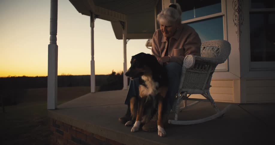 Elderly woman sitting on rocking chair on porch at sunrise petting and hugging her dog