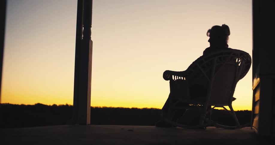 A woman sits on her porch at sunset as her dog gets up and walks away