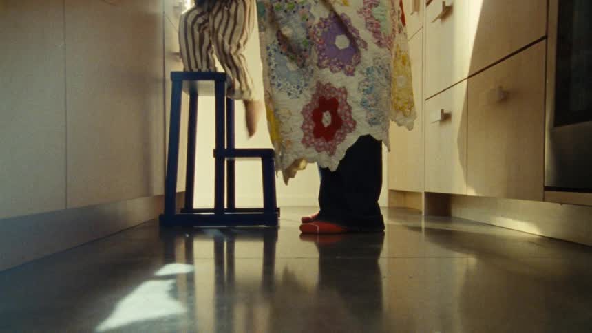 A lowangle shot in a kitchen showing a child on a step stool being covered with a patterned blanket by an adult