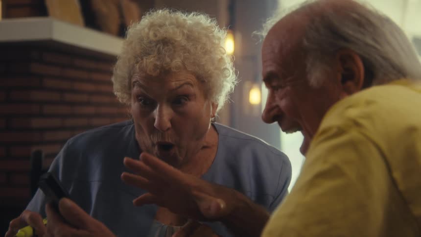 A closeup shot of an elderly couple with a woman with curly white hair and a man in a yellow shirt holding a smartphone and the woman reacts with surprise and then smiles joyfully