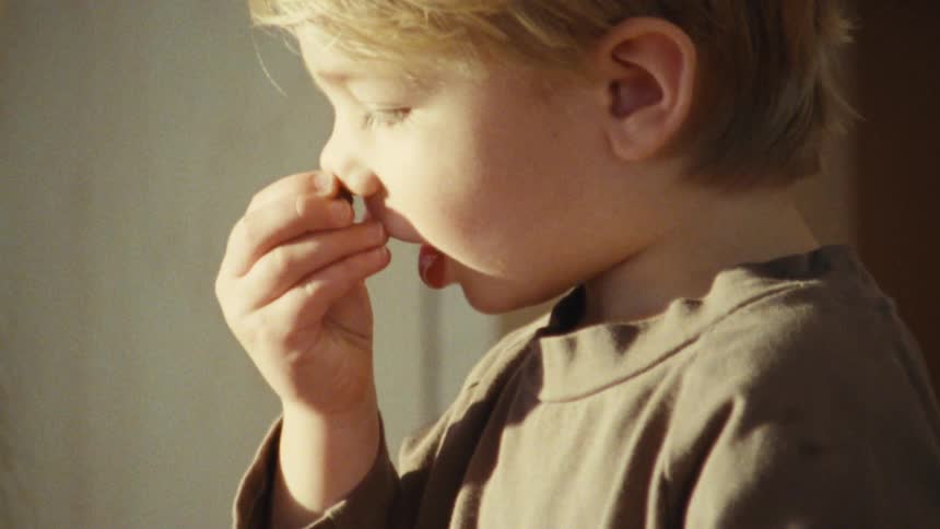 A child picks up a rock and holds it to his nose to smell it while his mother watches and laughs outdoors at midday
