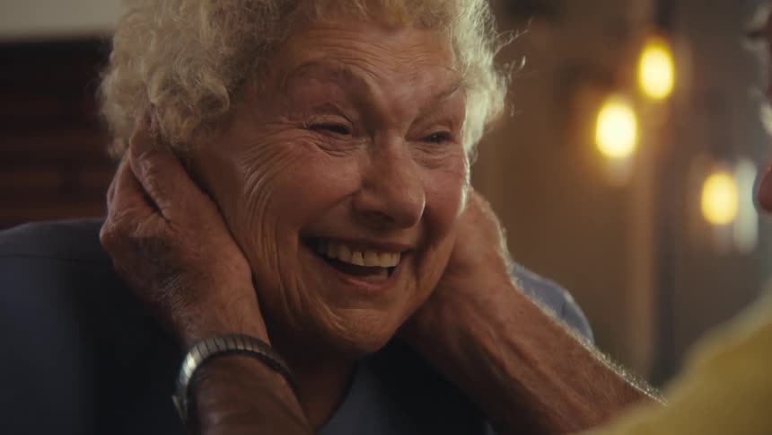 A happy elderly woman with curly white hair smiling with the hands of a man on her face