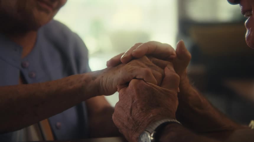 A close view shows an elderly mans hands clasped with an elderly womans hands on a table