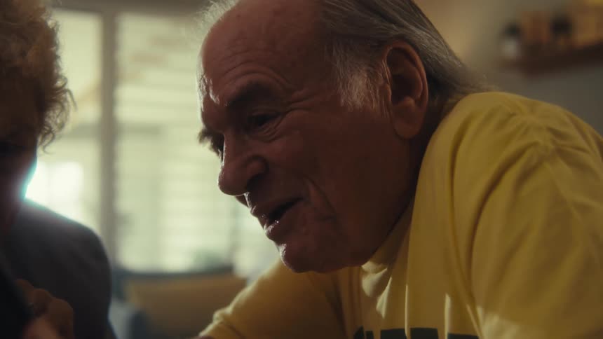 A man with white hair and a yellow shirt sits across a table from a woman with curly white hair as they talk and he smiles