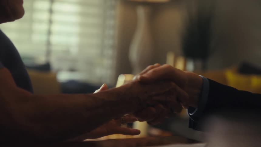An elderly woman shakes a man who is putting on a suit both seated at the dining table indoors at midday