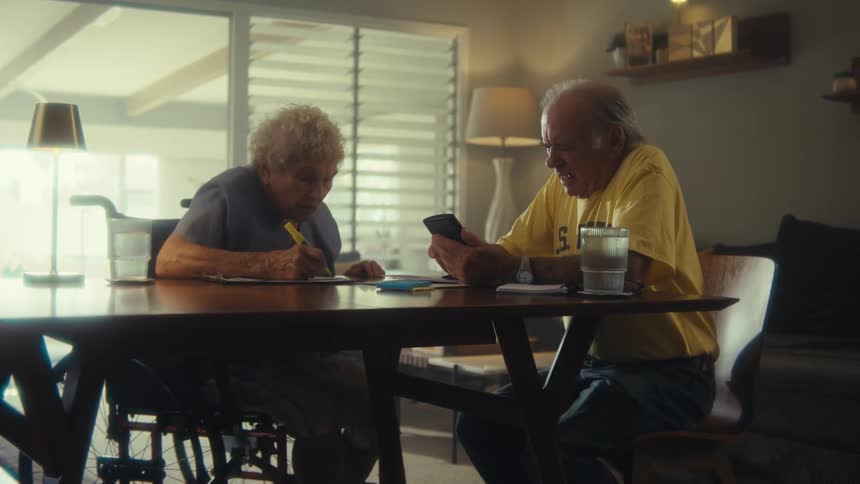 Elderly couple talks at a dining table with notes and a phone