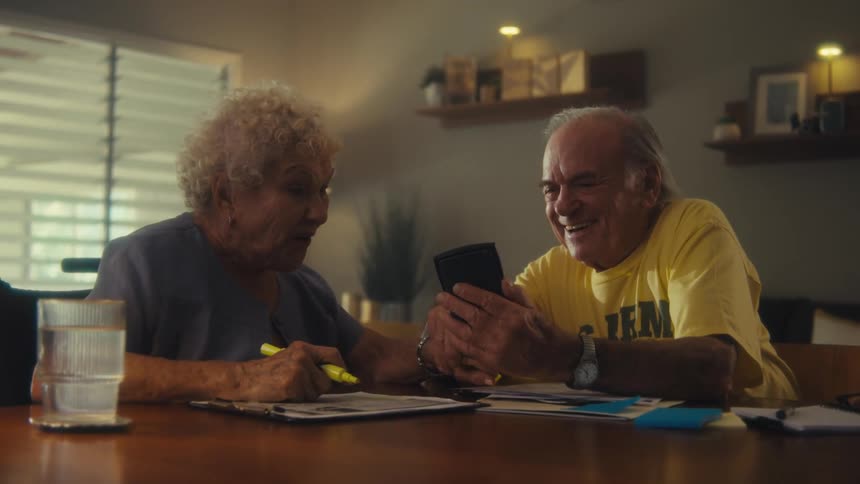An elderly couple sits at a table with a man looking at a phone and the woman with a newspaper both talking and laughing