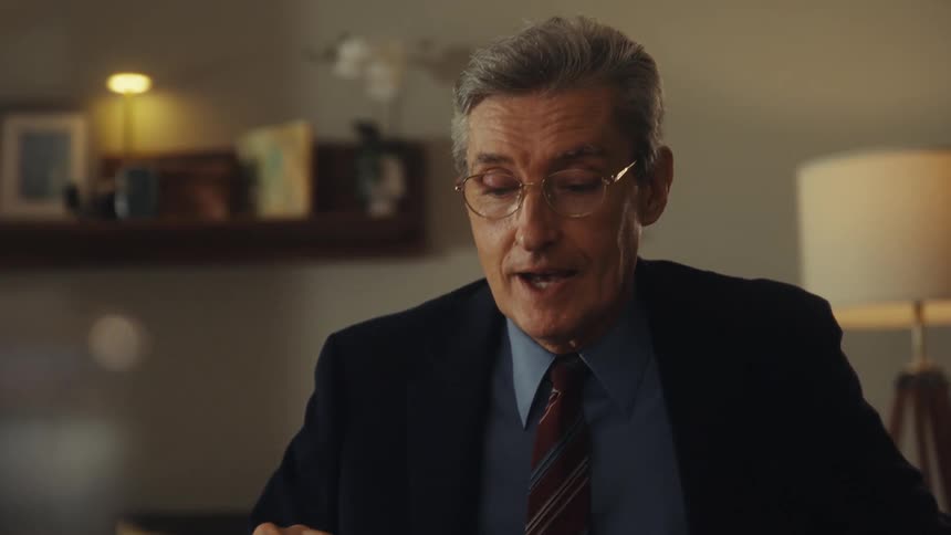 A closeup shot of a middleaged man with grey hair and glasses wearing a suit and tie smiling and gesturing as he speaks