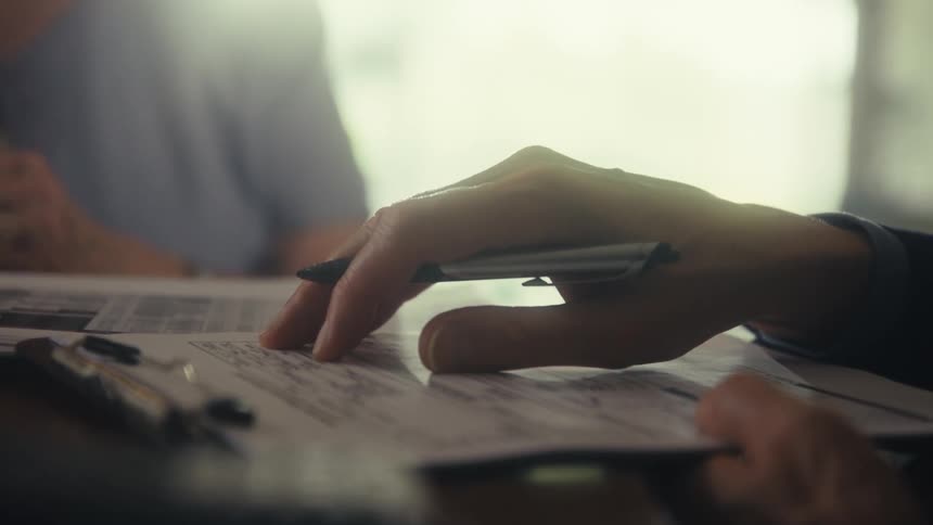 A close view shows a persons hand writing on a medical form on a clipboard with a black pen