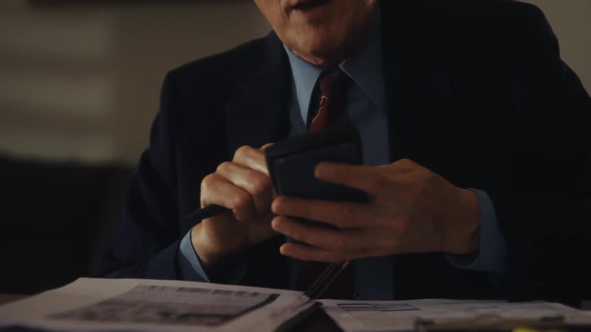 A middleaged man wearing a suit tie and glasses sits behind a desk with several documents on it he holds a pen while picking up his phone and scrolling through it as he talks to someone across from him