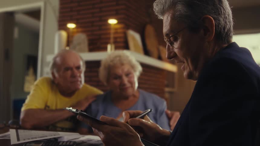 An elderly couple sits at a dining table watching a middleaged man at the other end wearing a suit writing on a notepad with a pen as he talks