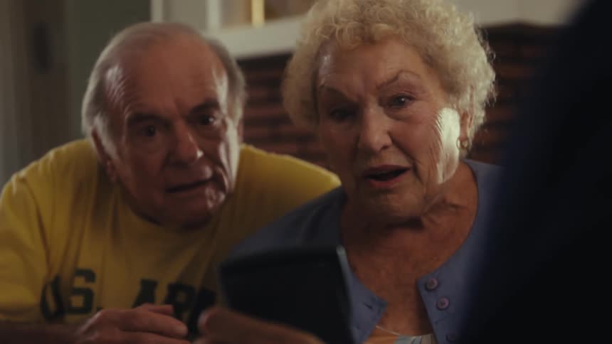 A man sits with an elderly couple punching numbers on a calculator and showing them the result the couple looks shocked and surprised then suddenly becomes excited and starts laughing