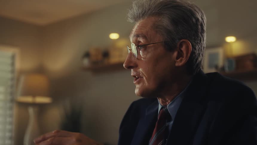 A man wearing a suit and glasses is seated at a table speaking and gesturing with his hands