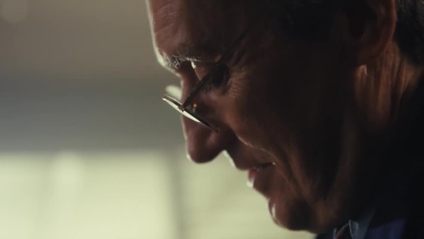 The video shows a man in profile wearing glasses smiling and talking the scene is lit from the side with some light reflecting off his glasses