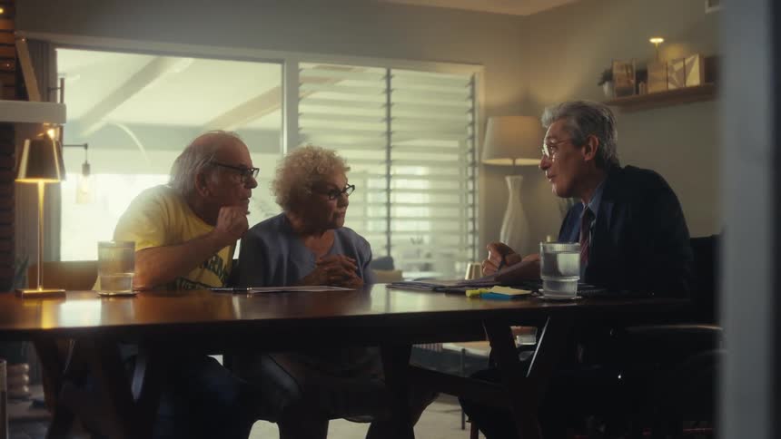 Two elderly individuals in a home setting discussing paperwork with a professional