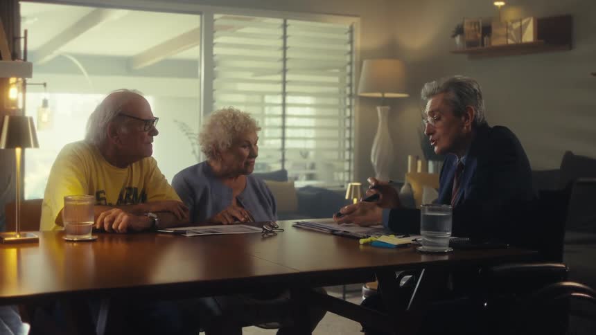 Three older adults are sitting around a table in what appears to be a living room or office two are a man and a woman sitting sidebyside listening to another man in a suit who is gesturing and speaking the man in the suit is holding a folder or book