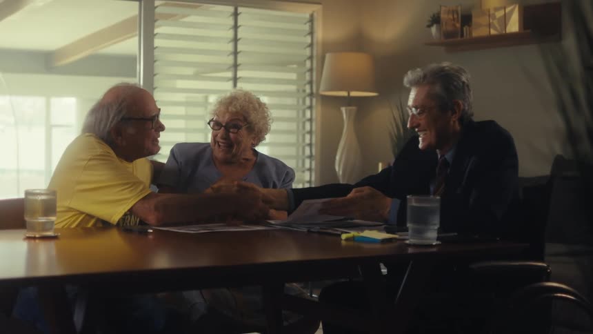A man in formal attire talking to an elderly couple and handing them a document with the couple happy and celebrating