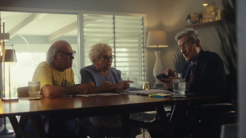 An elderly couple talking to an older man in formal attire who is explaining something to them and showing his phone and some documents