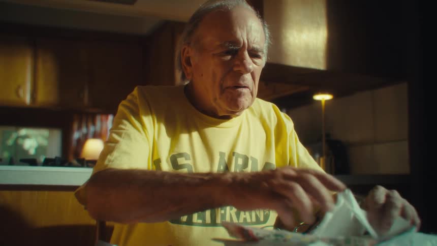 An elderly man wearing a yellow shirt is sitting at a kitchen table looking down and sorting through a pile of papers