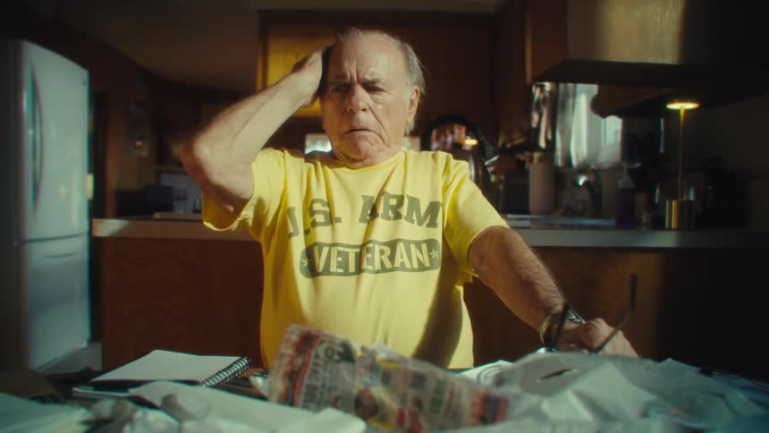 Elderly man in a yellow us army shirt appears in a kitchen looking serious while leaning on a table full of papers