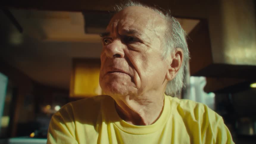Elderly man in a yellow tshirt looks to the side with a serious expression in a sunlit kitchen