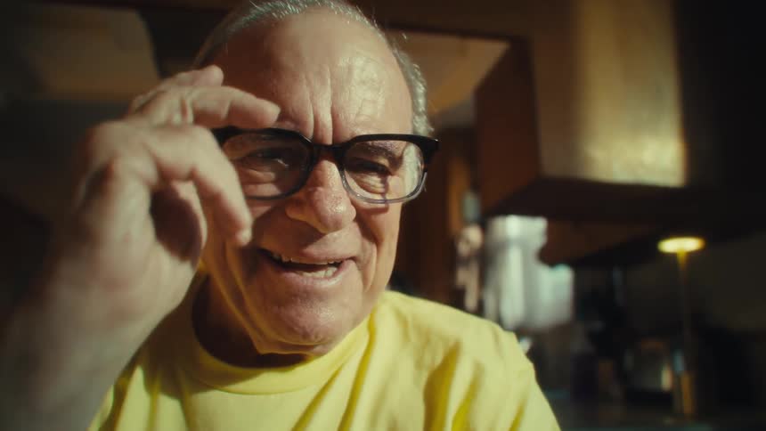 An elderly man wearing a yellow tshirt is smiling and putting on his glasses while he is looking to a smartphone screen