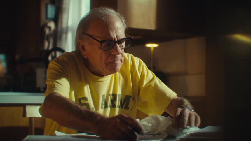 A man wearing a yellow us army tshirt and glasses sits at a table looking down at some papers and flipping through them with his hands