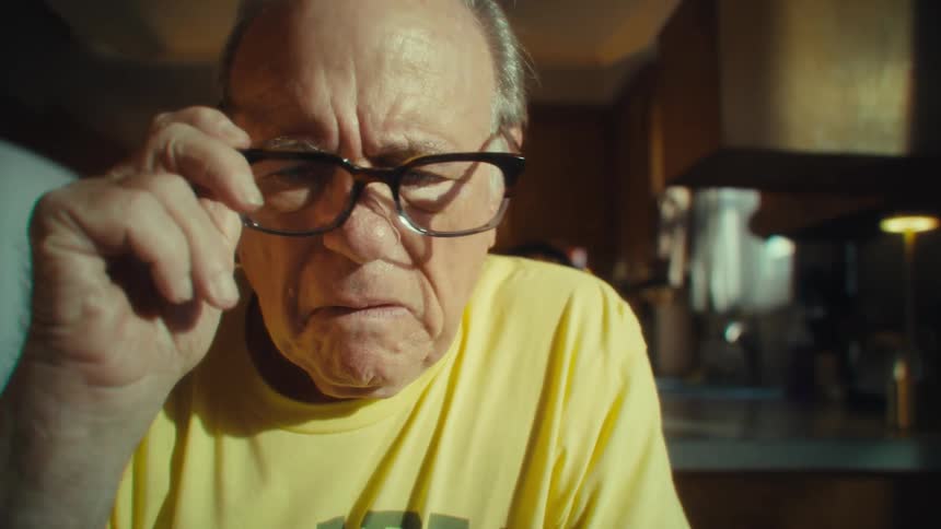 A man wearing a yellow shirt and glasses looks down at his phone then takes off his glasses and looks straight ahead with a sad expression on his face