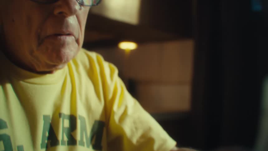 A man wearing a yellow shirt and glasses puts on his glasses and looks down at a handful of receipts