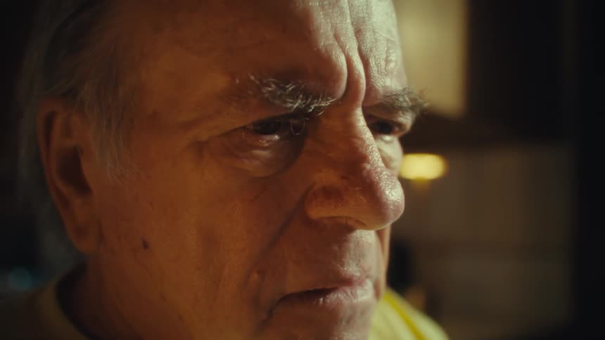 An elderly man wearing a yellow tshirt that says us army veteran sits at his dining table with several documents in front of him looking worried as he puts on his glasses